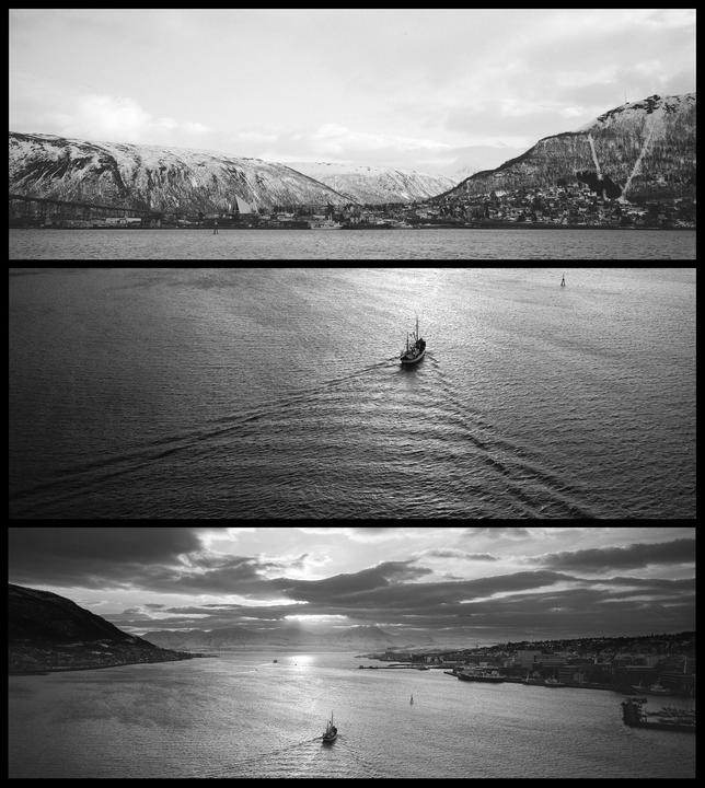 A boat leaves a wake in the water as it travels through a fjord surrounded by snow-capped mountains and a city.