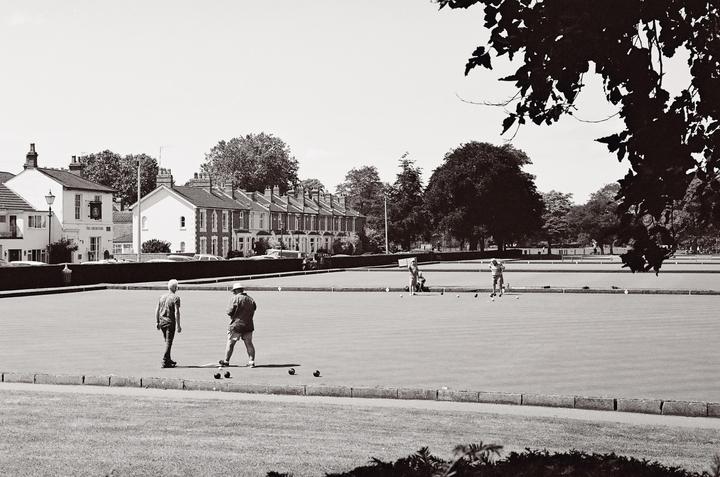 Several people are playing lawn bowls on a green field with houses and trees in the background.