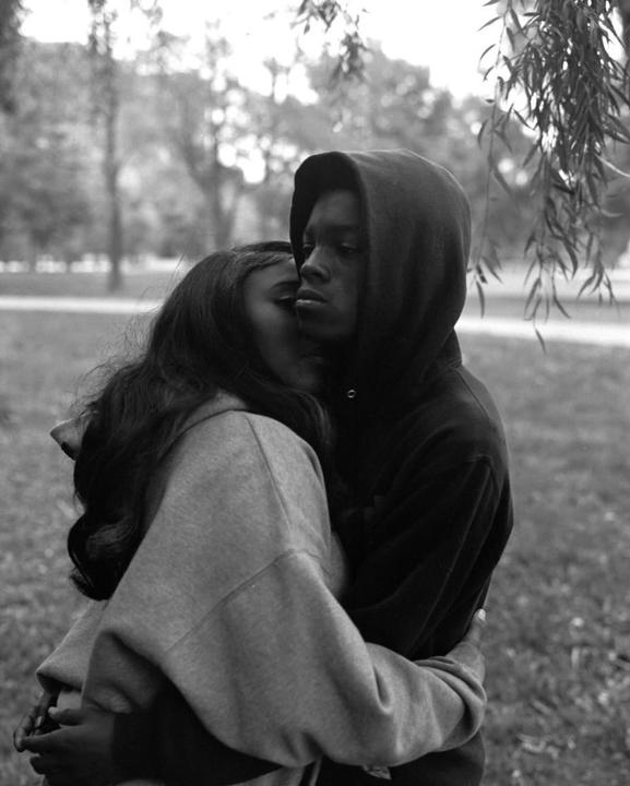 A couple embraces outdoors, with a man in a hoodie looking away as a woman rests her head on his shoulder.