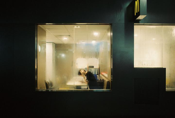 A person sits with their head in their hands inside a brightly lit fast food restaurant at night, viewed through a window.