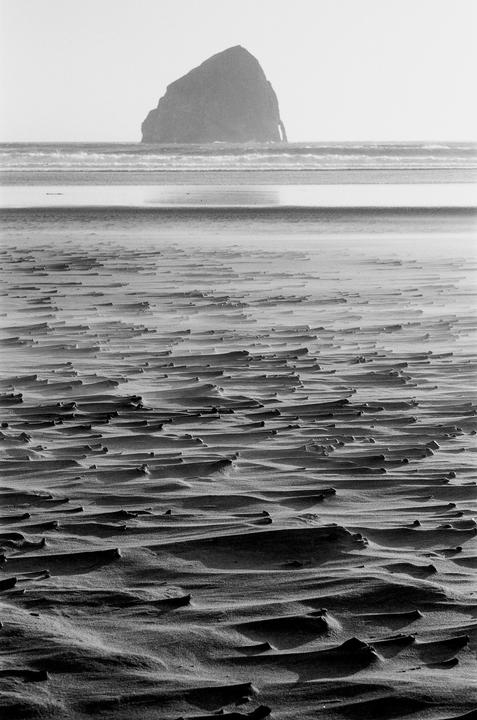 Wind blows sand across a beach with a large rock formation in the distance.
