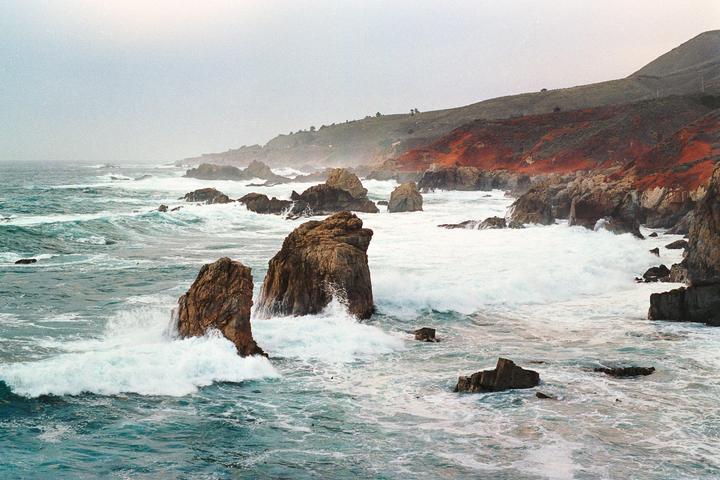 Waves crash against large rocks along a rugged coastline with a hillside in the background.