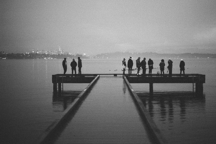 A group of people stand on a pier overlooking a body of water with a city skyline in the distance.
