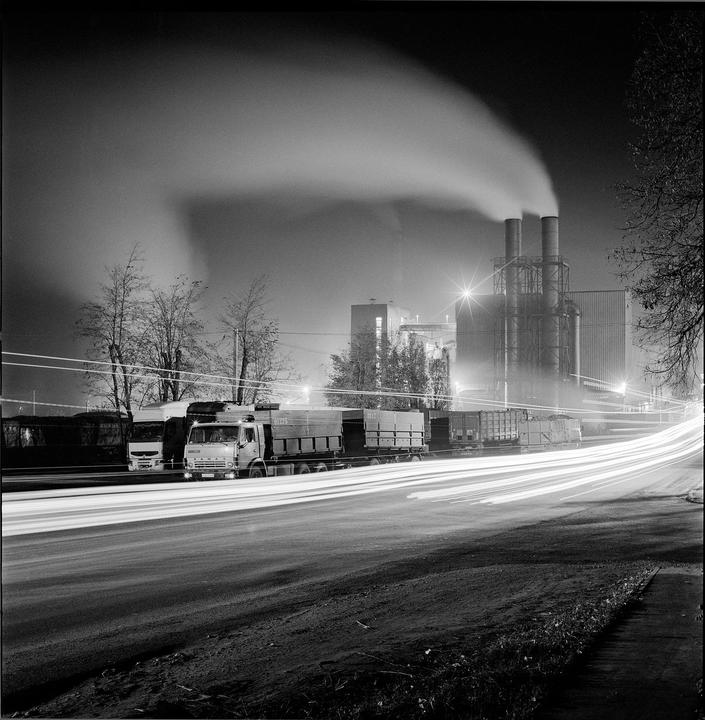 A sugar factory with smoke billowing from its chimneys stands illuminated at night, with light trails from passing vehicles on a nearby road.