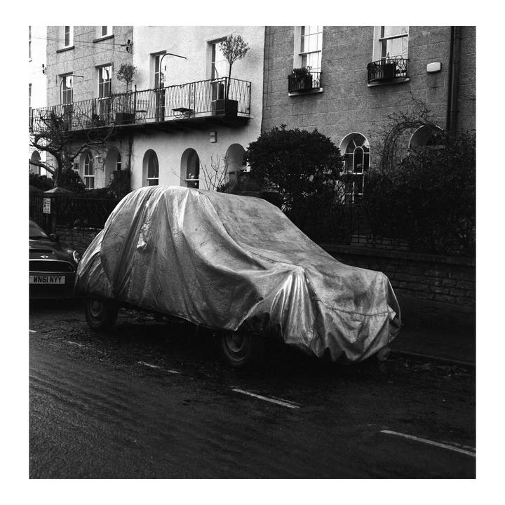 A car covered with a tarp is parked on a wet street in front of a building with balconies.