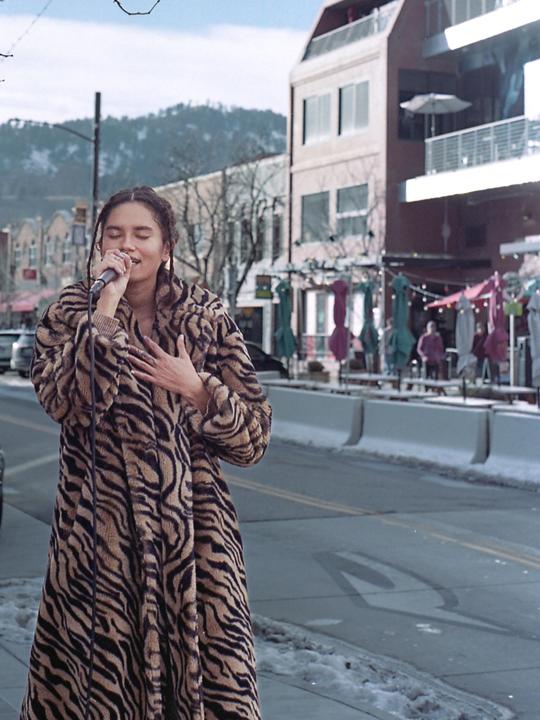 A woman in a tiger print coat sings into a microphone on a street with snow on the ground and mountains in the background.