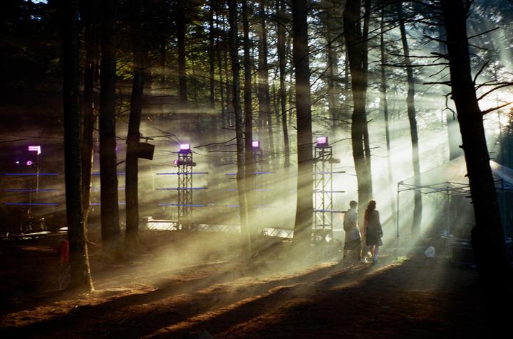 Two people stand in a hazy forest with sunbeams shining through the trees and stage equipment in the background.