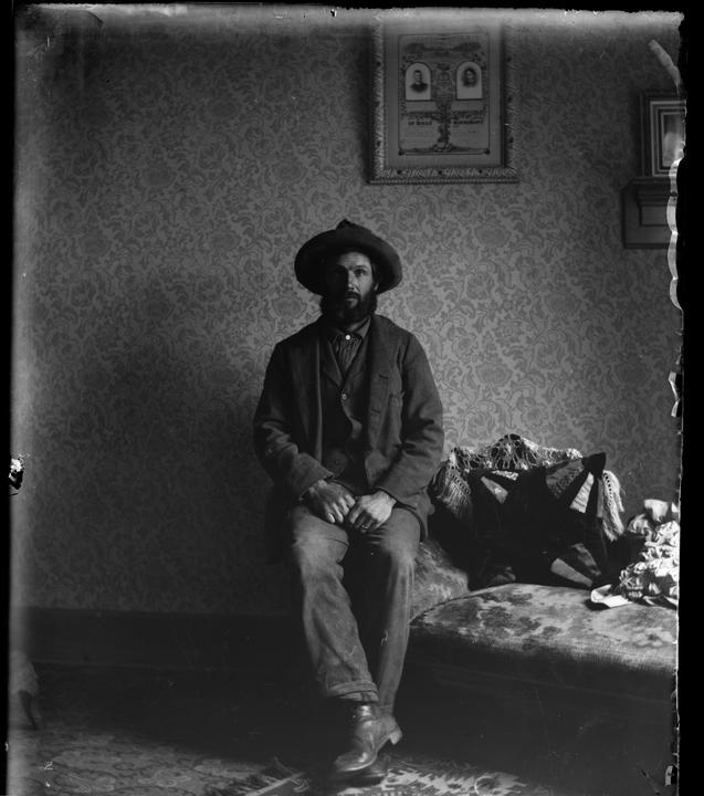 A man with a beard and a hat sits on a chaise lounge in a room with patterned wallpaper.