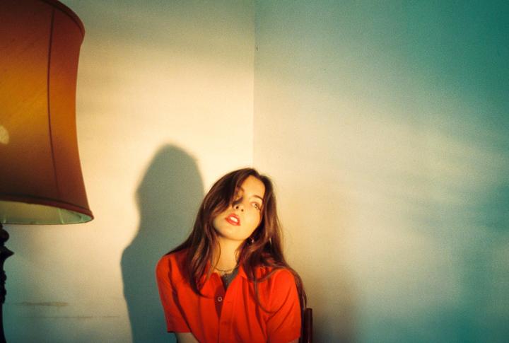 A woman in a red shirt sits in a corner with a lamp to her left, casting a shadow on the wall.
