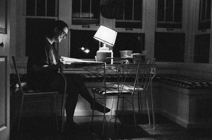 A woman with glasses sits at a table with a lamp, reading or working on something.