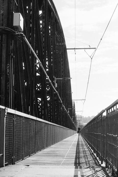 A person walks along a long, narrow bridge with a metal truss structure on one side and a metal fence on the other.
