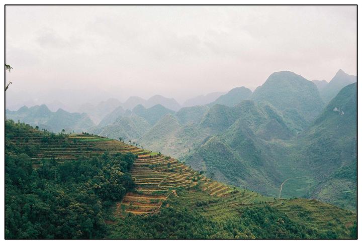 Terraced fields and lush green mountains stretch into the hazy distance under a cloudy sky.