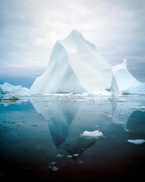 A large iceberg is reflected in the dark water, surrounded by smaller pieces of ice.