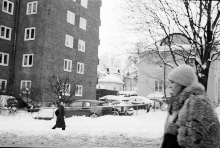 A woman in a fur coat and hat walks past a snow-covered street with buildings and cars.