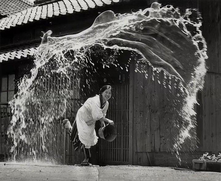 A woman in traditional clothing smiles while being splashed by a large volume of water in front of a wooden building.