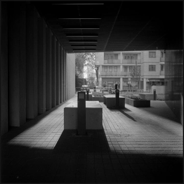Sunlight casts strong shadows on a tiled walkway next to a building with a distant building in the background.