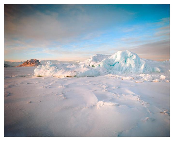A vast expanse of snow and ice covers the ground, with large ice formations in the middle ground and mountains in the distance under a blue sky.
