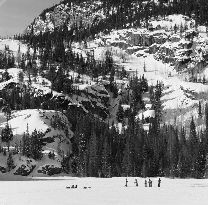 A group of people stand on a frozen lake surrounded by snow-covered mountains and pine trees.