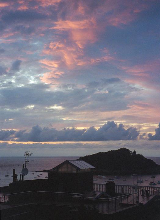 A dramatic sunset with pink and purple clouds hangs over a bay with boats and a dark island.
