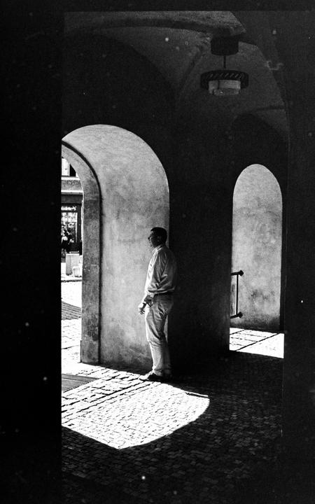 A man stands in an arched passageway with a patch of sunlight on the cobblestone ground.