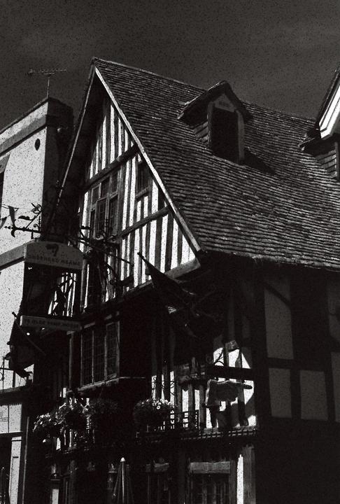 A black and white photo shows a timber-framed building with a steep, tiled roof and a dormer window.