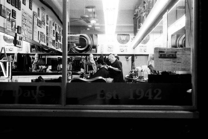 A barber cuts a man's hair in a barbershop seen through a window.