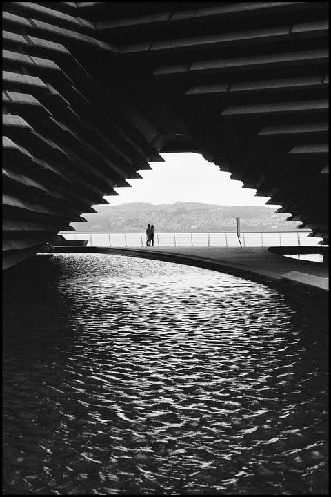 A couple stands by a river with a distant hillside under a large, dark, geometric structure.
