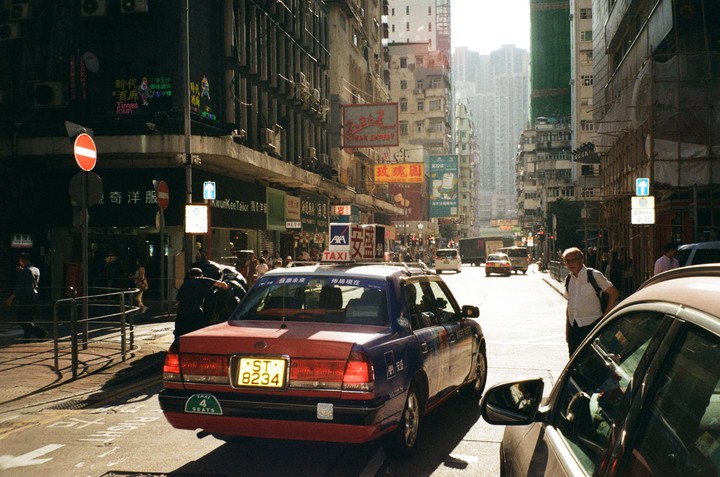 A red and blue taxi is parked on a busy street in Hong Kong with tall buildings and many signs in the background.