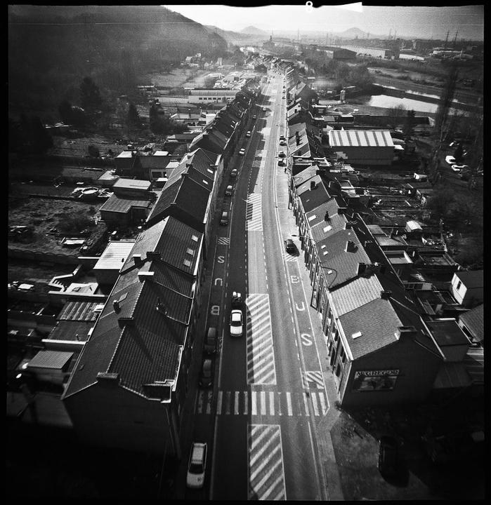An aerial view shows a long street lined with houses and cars, with mountains in the distance.