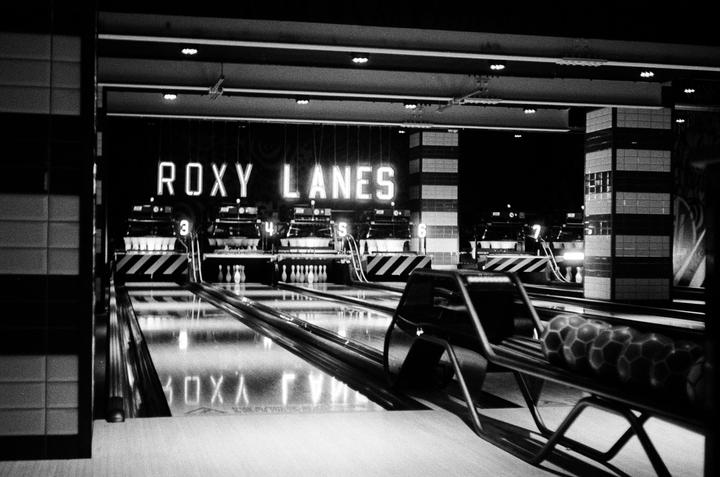 A black and white image shows a bowling alley with the neon sign "ROXY LANES" above the lanes.