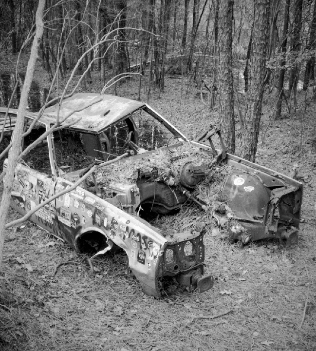 A heavily damaged car chassis covered in stickers sits abandoned in a forest.