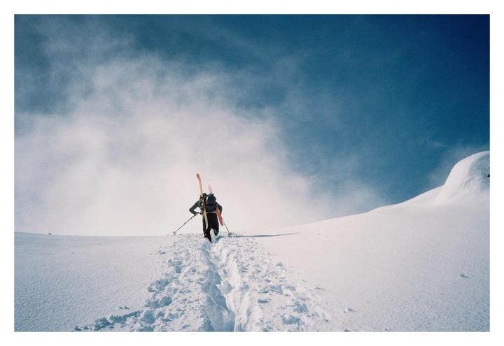 A person with skis and poles walks up a snowy mountain, leaving footprints in the snow.