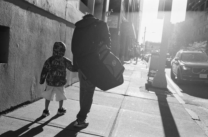 A man and a child walk down a sunlit street, holding hands.