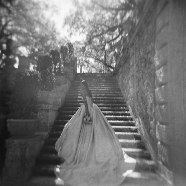 A woman in a wedding dress stands on a stone staircase with trees in the background.