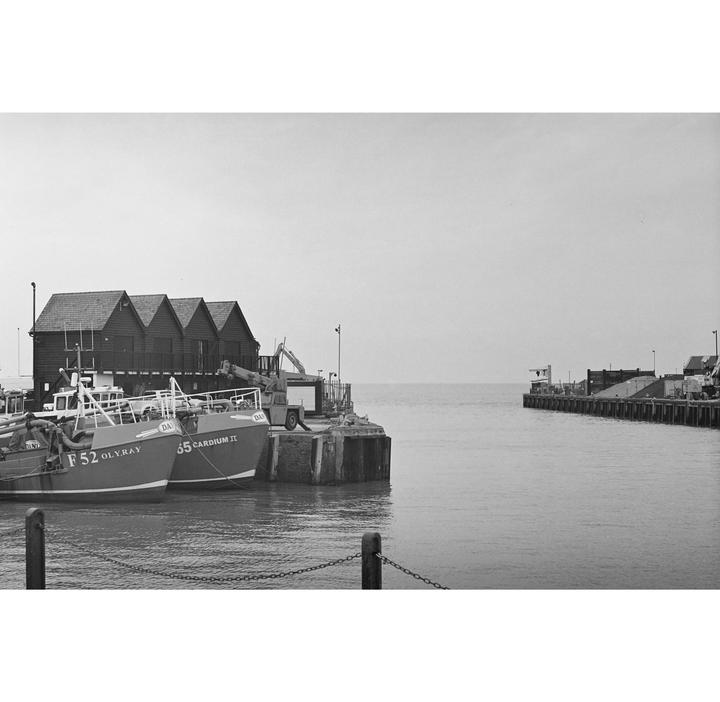 Two boats are docked next to a pier with three small buildings and a crane.