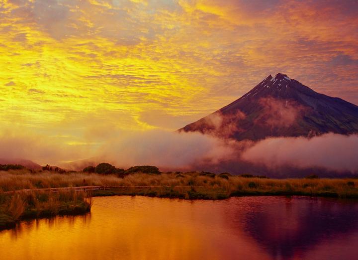 A mountain peak rises above a body of water with a vibrant orange and yellow sky at sunrise.