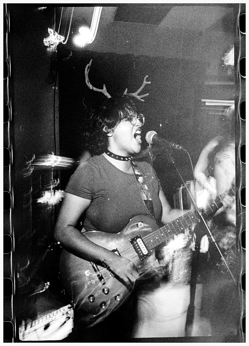 A woman wearing antlers and glasses sings into a microphone while playing an electric guitar on a dark stage.