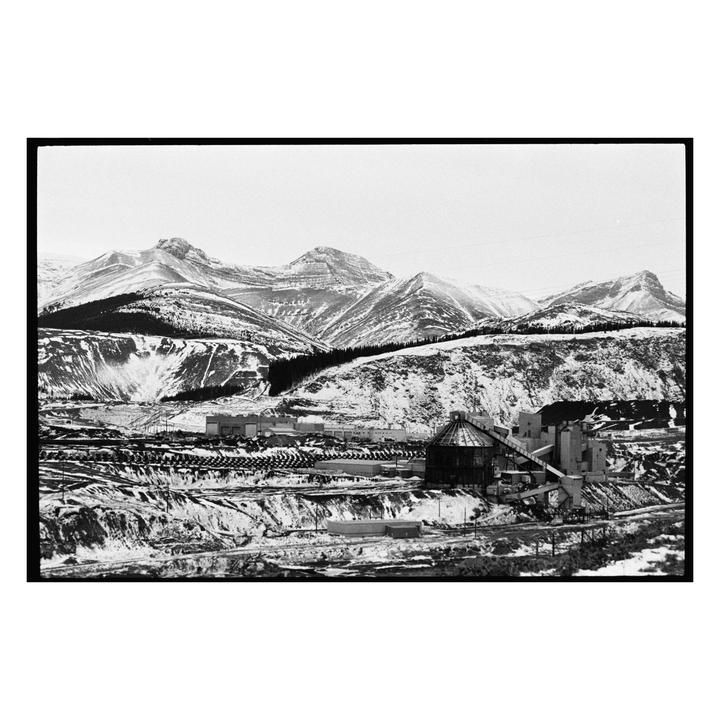 A large industrial facility with silos and conveyor belts sits in a snowy valley with mountains in the background.