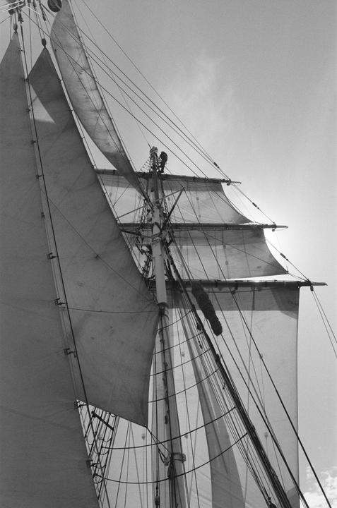 Two men are visible high up on the mast of a sailboat with many sails and ropes.