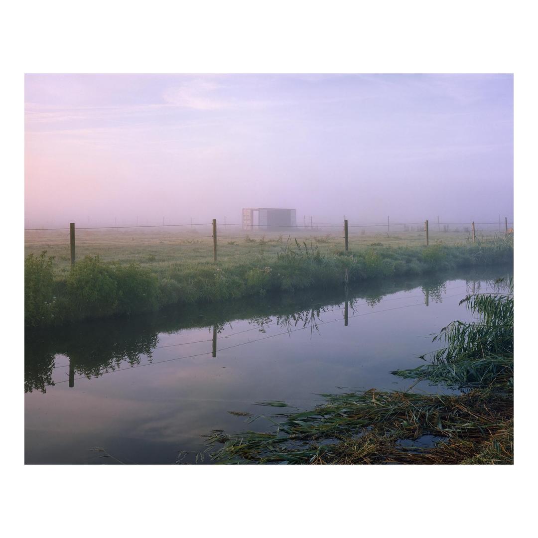 A misty field with a fence and a canal in the foreground, and a building in the distance.