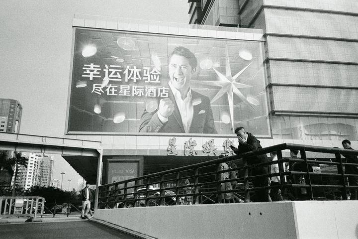 A large billboard with a man in a suit smiling and clenching his fist is above a street with people and buildings.