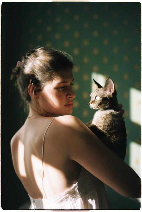 A woman holds a cat in her arms, looking at it with soft light falling on her back and the cat.