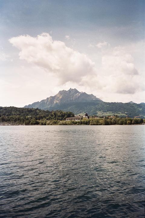 A body of water with a forested shore and buildings in the distance is overlooked by a large mountain under a cloudy sky.