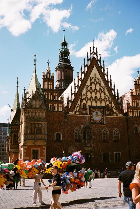 Two people carry large bundles of character balloons in a cobblestone square in front of a historic brick building with a clock tower.
