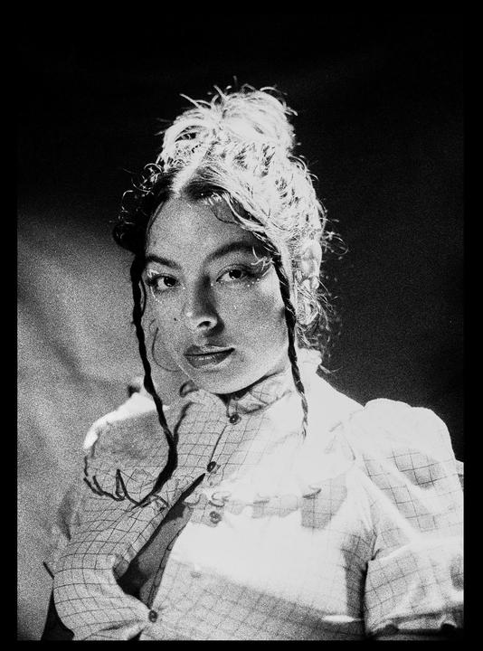 A woman with her hair in a bun and two braids looks at the viewer.