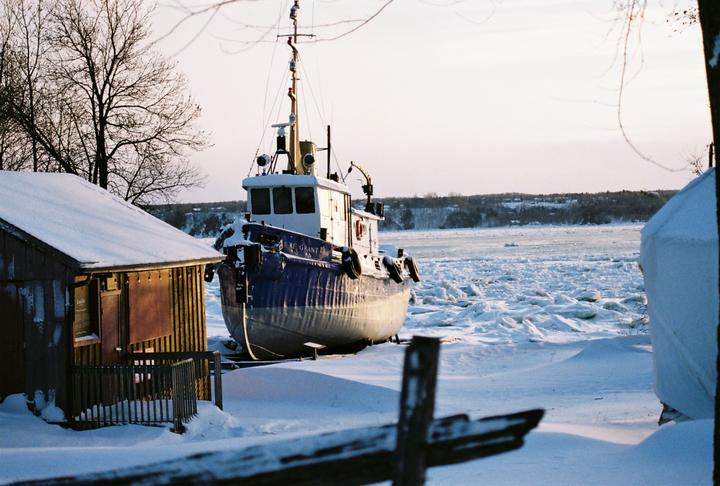 A blue and white boat sits on a snowy bank next to a frozen body of water with a small wooden building in the foreground.