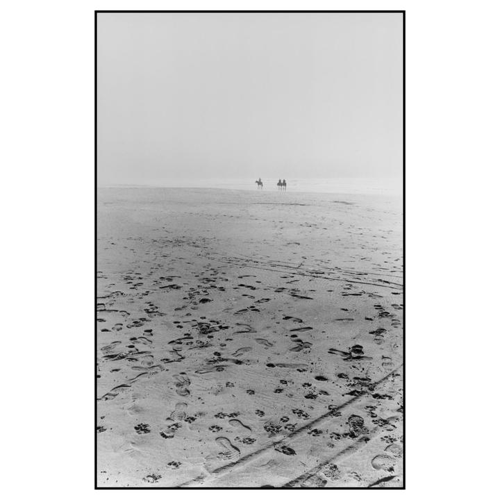 Two people on horseback are visible in the distance on a hazy beach covered in footprints.