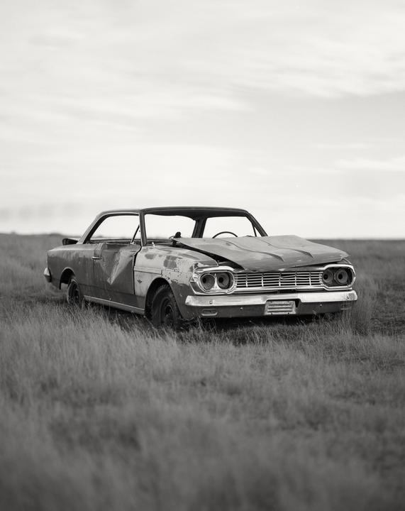 A wrecked car sits in a field of tall grass under a cloudy sky.