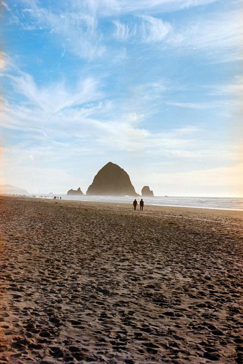 Two people walk on a sandy beach toward a large rock formation in the distance under a blue sky with wispy clouds.