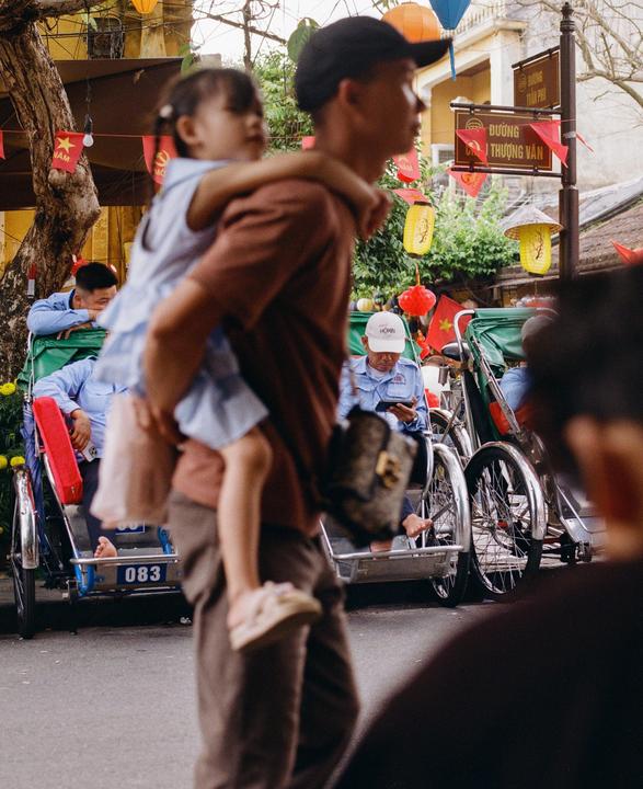 A man carries a child on his back while walking past cyclos and people on a street decorated with lanterns and flags.